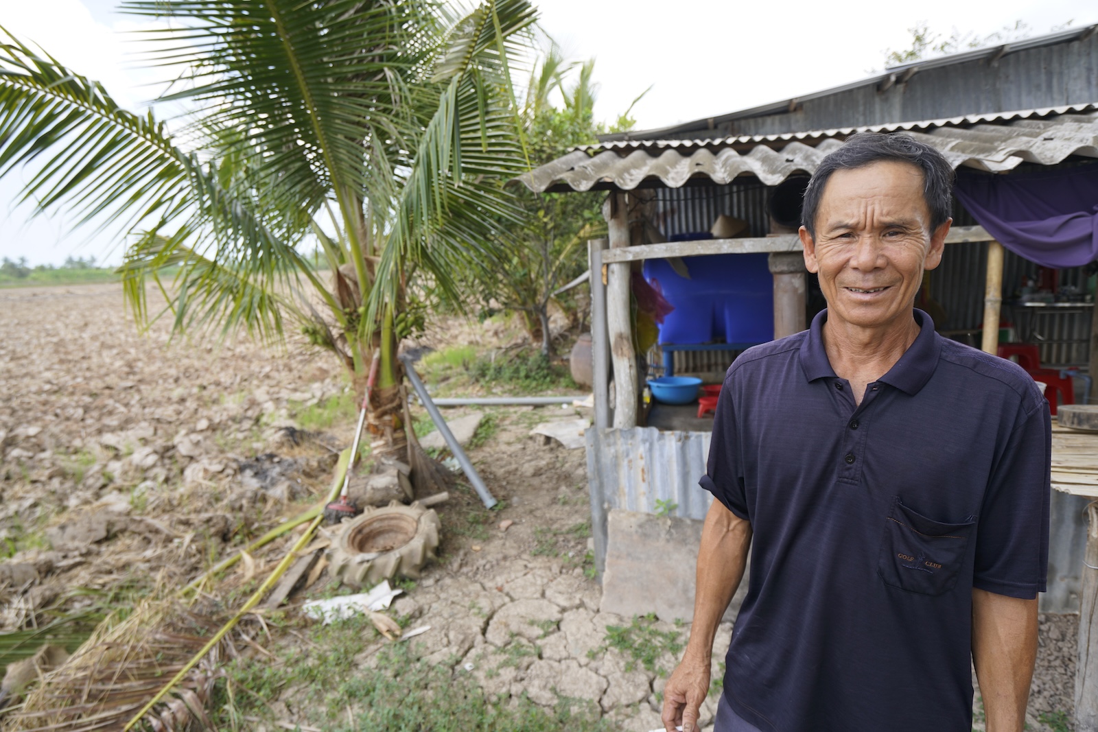 A farmer stands beside a field of cracked, dried earth next to his home