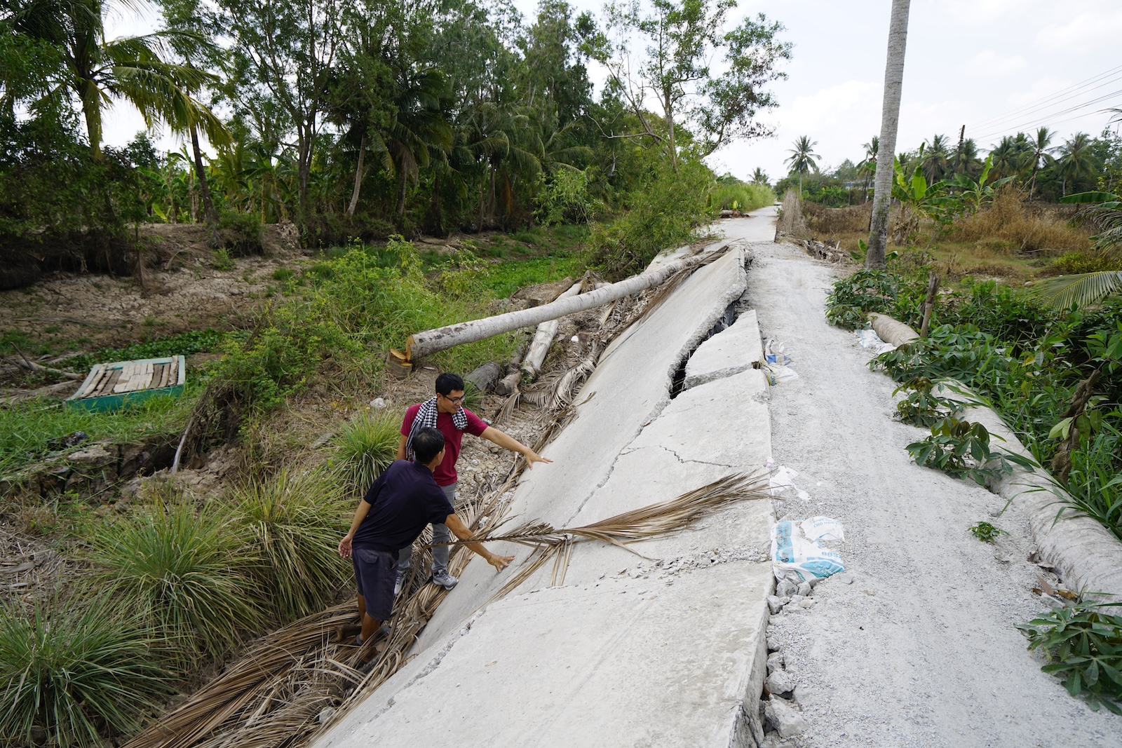 Two men inspect a rural concrete road that has split and slumped toward a canal